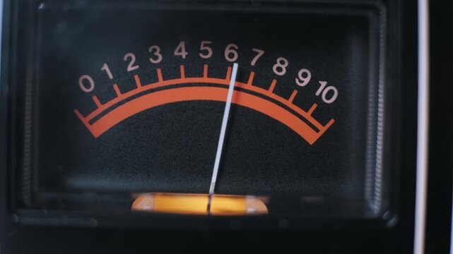 Macro shot of an analog VU meter with a needle fluctuating across a red numeric scale. The vintage equipment features a grainy display measuring audio signal levels.