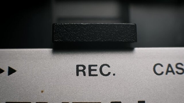 Extreme close-up macro view of a male finger pressing the black textured REC button on a silver vintage player. The panel shows printed text activating the recording function.