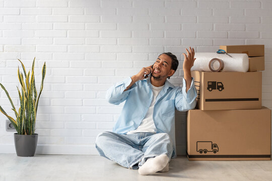 Young man sitting on floor talking on phone next to cardboard moving boxes, bubble wrap and tape. Moving day concept, real estate, relocation