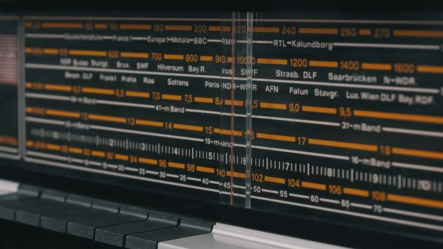 Close-up pan of a vintage radio tuner analog display with orange markings. The scale shows wavebands and European city station names on a black background.