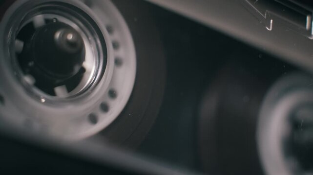 Angled macro shot of a classic audio cassette playing with a transparent reel. Red markings spin as dark magnetic tape unwinds inside the player in an analog setup.