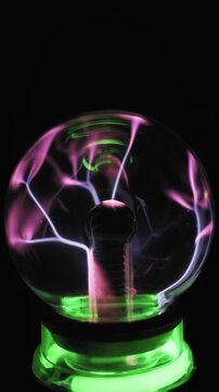 Vertical angled view of a plasma globe with pink and green bolts from the central electrode. The glass sphere features ionized gas tendrils moving electric currents on a dark backdrop.
