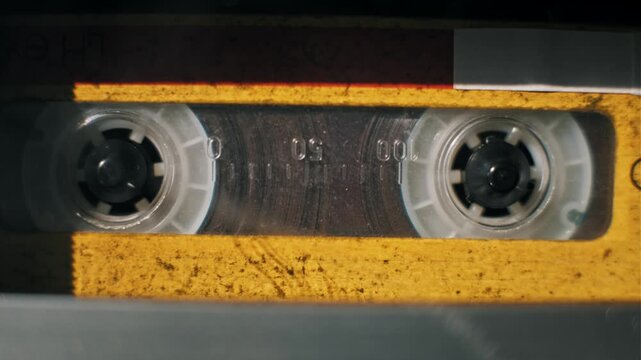 Close-up of a hand opening a vintage tape deck and inserting an old yellow audio cassette. The player mechanism engages as reels spin under sunlight and moving shadows.
