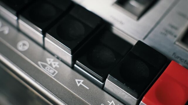 Macro shot of a finger pressing the black play button on a vintage cassette tape recorder. The metal deck features control panel symbols and keys for retro audio.