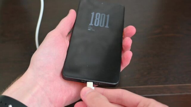Human hand holding black smartphone with charging cable on wooden desk close up