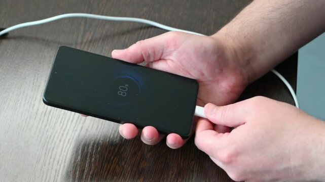 Human hand holding black smartphone with charging cable on wooden desk close up