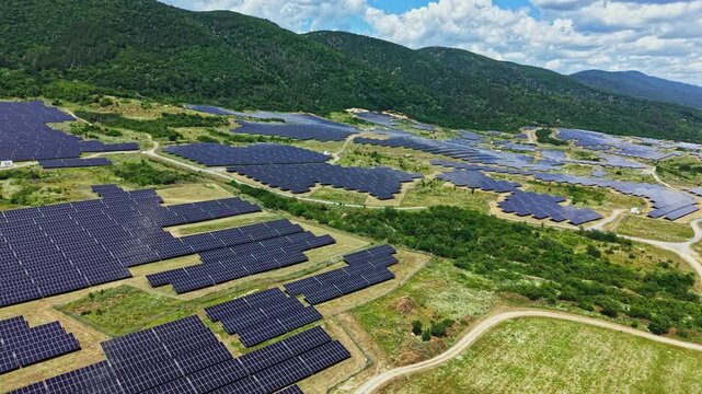 Rows of solar panels cover the ground in a vast area near hills. The sky is blue with some clouds. This site shows the use of solar energy for power generation.
