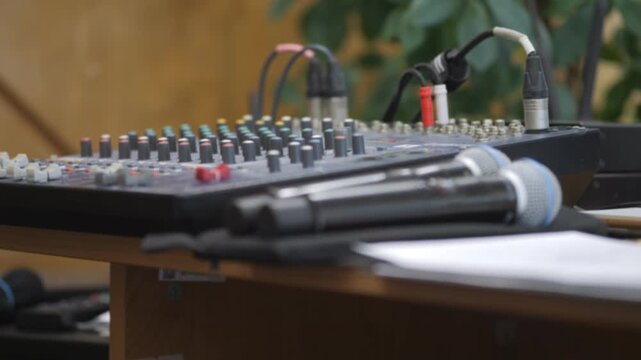 Audio mixer and microphones sit on wooden table. Cables connect devices to mixing board. Background shows green plants and warm lighting. Ideal for music production, live events, or studio setups