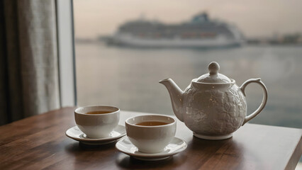 Fototapeta premium Two cups of tea and a ceramic teapot on a wooden table with a blurred cruise ship sailing on the sea in the background