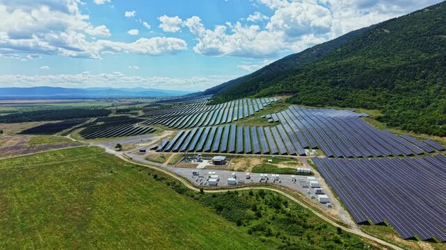 A solar farm with many rows of solar panels is placed on a hillside. The area is sunny and the landscape includes fields and mountains. White storage units are present nearby.