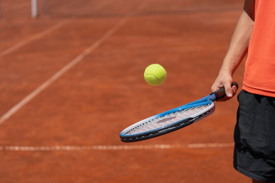 Teenage male tennis player balancing tennis ball on racket on clay court. Horizontal composition with copy space for sports lifestyle concept, training and healthy active youth