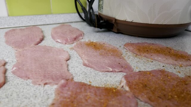 Raw, thin pork cutlets are arranged on a kitchen countertop, some seasoned with a dry spice rub. Close-up shot pans slowly across the meat, showing the texture of the raw pork and the spices.