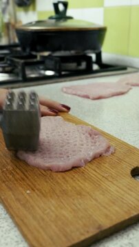 Woman's hand uses a metal meat mallet to tenderize a raw pork fillet on a wooden cutting board. Close-up shot captures the food preparation process with a stove and pan in the background.