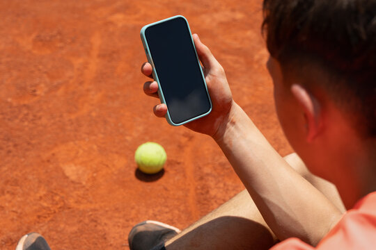 Junior tennis player holding smartphone with blank screen on clay court. Mobile app mockup and empty court provide copy space for app, advertising, sports communication and digital technology concepts