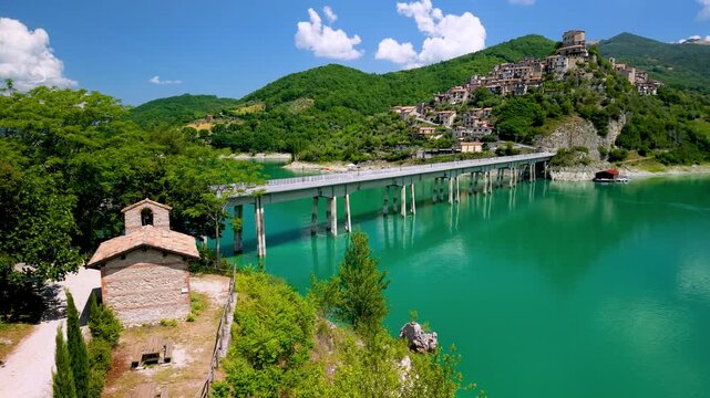  Castel di Tora Turano Lake Italy 4K Drone footage. Diagonal bridge on stilts over turquoise water to hilltop medieval borgo town with church. Borghi piu belli d Italia most beautiful villages