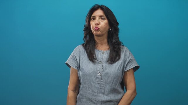 Woman pouting lips in a gray blouse against a teal studio backdrop, standing centered and facing camera; playful confidence.