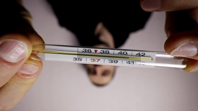 Male hands hold a glass mercury thermometer showing a dangerously high temperature of 41 degrees Celsius. A close-up view with a concerned face reflected in the background.