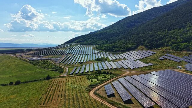 Solar panels are placed in long rows on flat land in a valley. The hills rise in the background under a bright sky with clouds. The scene shows a renewable energy setup.