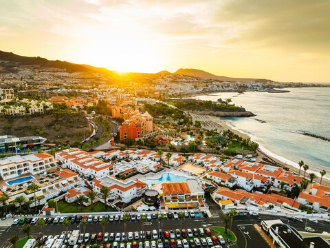 Aerial view of resort hotels with swimming pools, palm trees, and a sandy beach along the coastline during sunset in Costa Adeje, Canary Islands, Spain.