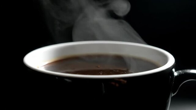 Hot steaming cup of fresh black coffee or dark espresso served in a ceramic mug against a dramatic solid black background with visible white vapor