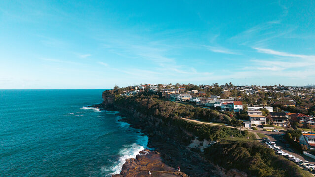 Aerial view of the coastal cliffs and residential area featuring houses overlooking the Pacific Ocean, rocky shoreline, and clear blue sky in Dee Why, New South Wales, Australia.