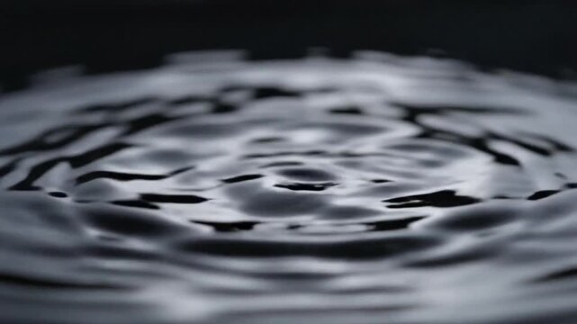 Macro shot of dark water surface with multiple concentric ripples and falling droplets creating dynamic shapes