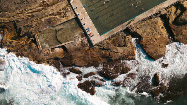 Aerial view of Dee Why Rock Pool with people swimming on the concrete edge, surrounded by rugged coastal rocks and crashing ocean waves in Dee Why, New South Wales, Australia.