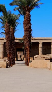 Seti I mortuary temple entrance with palm tree alley and ancient stone ruins in Luxor Egypt. Scenic pathway under vivid blue sky.