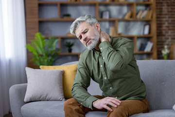 Fototapeta premium Middle eastern man experiencing neck pain and discomfort, touching his neck while sitting on a sofa at home, highlighting issues of health, stress, and lifestyle