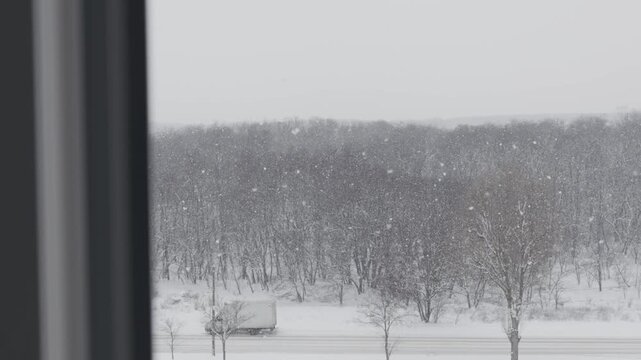 View from a window showing a truck and car driving on a road through a snowy forest during heavy snowfall. Concept of bad weather conditions, winter transportation, and scenic cold landscape.