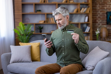 Fototapeta premium Mature man sitting on a sofa in his living room, looking puzzled and concerned while holding a credit card and checking his smartphone, experiencing online shopping problems or financial stress