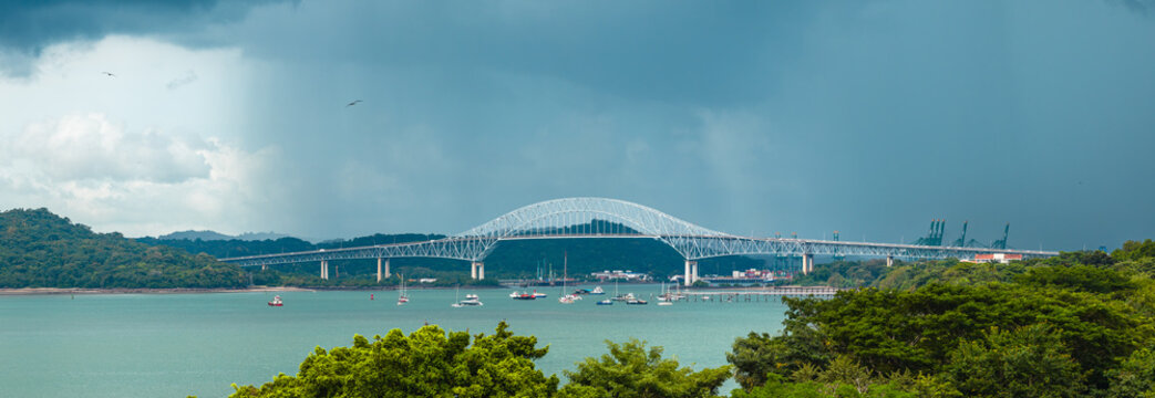 Aerial view of the Bridge of the Americas spanning the Panama Canal entrance under a dramatic stormy sky with lush vegetation and boats in the water in Panama, Provincia de Panama, Panama.