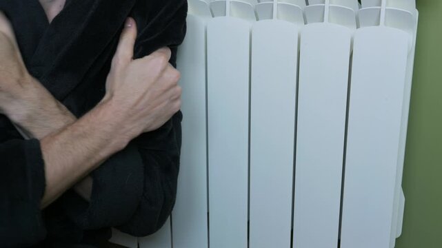 A frozen man in a black bathrobe shivers and hugs himself next to a white radiator. Concept of heating problems, rising energy costs, and freezing indoor temperatures in winter.
