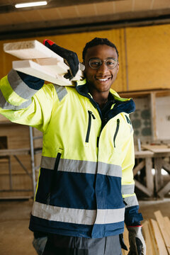 Portrait of smiling young vocational male student in high visibility safety jacket carrying wooden planks on shoulder