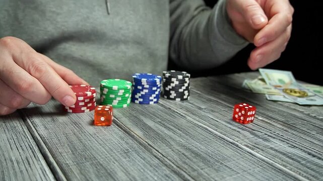 Person in gray sweater places poker chips and dice on a wooden table, with cash visible, showcasing a gaming setup in a dimly lit environment