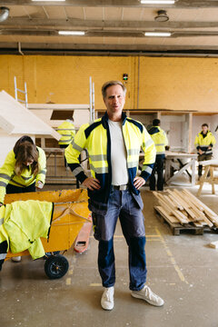 Full length portrait of male teacher wearing reflective jacket standing with arms akimbo in trade workshop