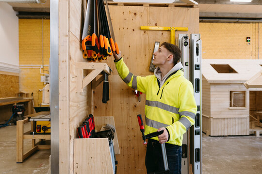 Young male student removing hand saw from tool rack while holding clamp at vocational workshop