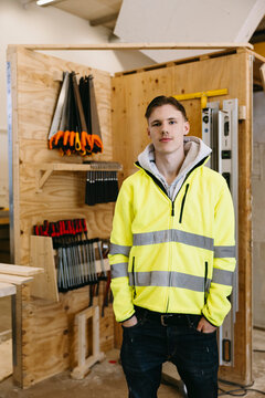 Portrait of young male student wearing reflective jacket standing with hands in pockets near tool rack at workshop