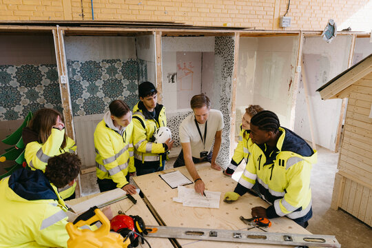 High angle view of instructor guiding students wearing reflective jackets around wooden work table inside construction t