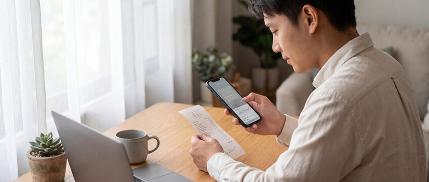 Scanning receipt with smartphone at a wooden desk with a laptop, coffee mug, and bright natural window light for digital expense tracking, personal budgeting, and tax management