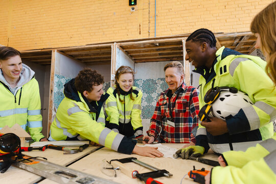 Smiling senior instructor in plaid shirt reviewing blueprint with vocational students around tool covered workbench at t