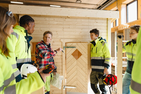 Senior instructor holding blueprint while pointing at wooden mock up house door while instructing vocational students in
