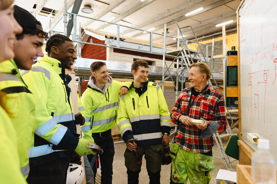 Smiling senior male professor talking with students while standing near whiteboard in workshop at vocational school