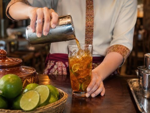 Thai-Eistee mit Limette Zubereitung &ndash; Kellnerin gie&szlig;t aromatischen Tee aus einem Shaker in ein Glas mit Eis und Zitrusscheiben. Warme Restaurantstimmung und authentische Bar-Szene.