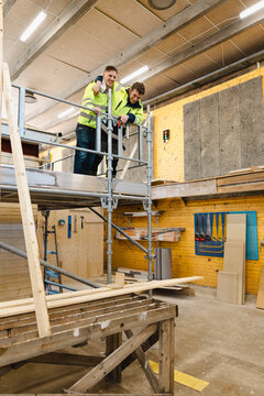 Smiling male vocational students standing on elevated scaffolding unit while enjoying leisure time inside carpentry work
