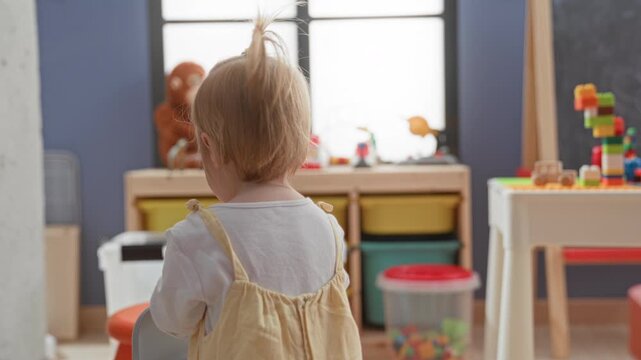 Toddler girl holding chair beside toy storage and stuffed monkey in play area inside a building; playful curiosity.