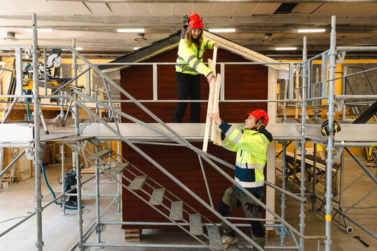 Smiling female student standing on scaffolding stairs and passing wooden planks to classmate working on roof framing of