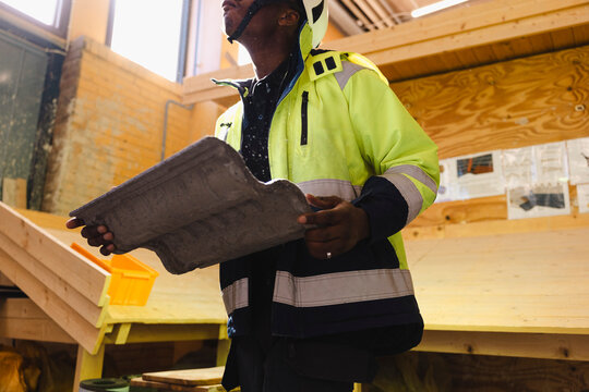 Low angle view of vocational male student wearing reflective jacket walking with concrete roofing tile inside building c