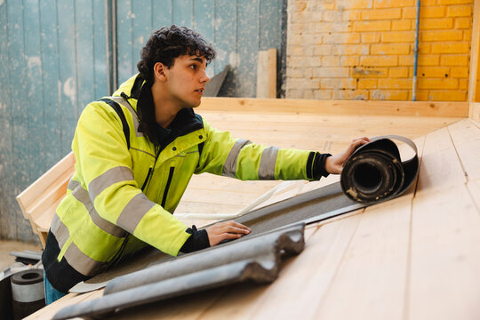 Male trainee wearing reflective jacket unrolling waterproof roofing underlayment on slanted wooden structure during trai