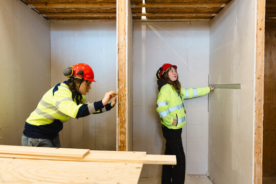 Female trainees wearing reflective jacket measuring drywall panel with ruler inside narrow construction booth at worksho